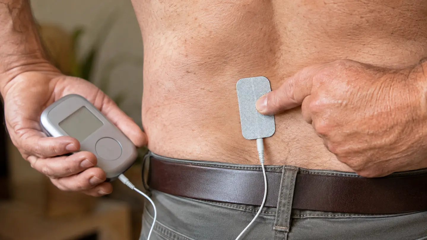 Close-up of a hand placing a TENS pad on a person's lower back just above the belt line